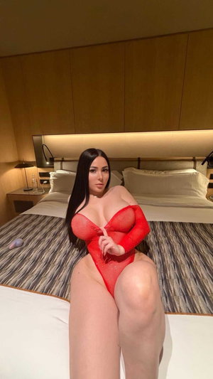 Buxom woman in red mesh lingerie on bedroom bed.