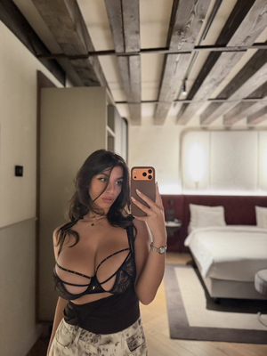 Woman in black lace bra and patterned pants takes mirror selfie in bedroom.
