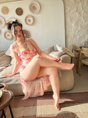 Woman in floral dress sits on couch, legs crossed, in cozy indoor setting.