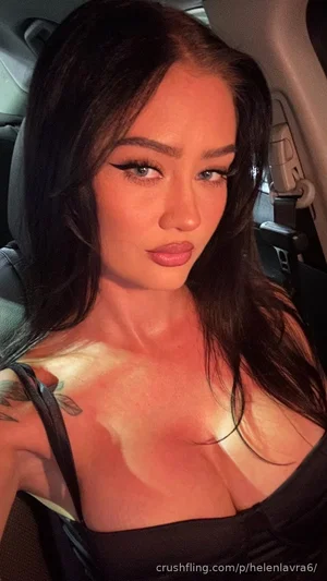 Woman with long dark hair and makeup wearing black top, taking selfie in car.