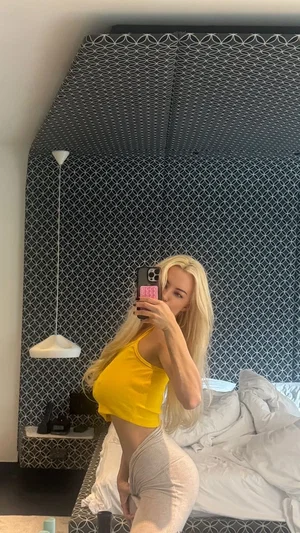 Blonde woman in yellow top and gray pants takes mirror selfie in bedroom.