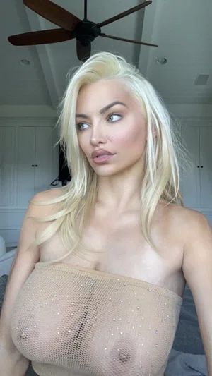 Blonde woman in sheer top with embellishments, indoors, looking to side.