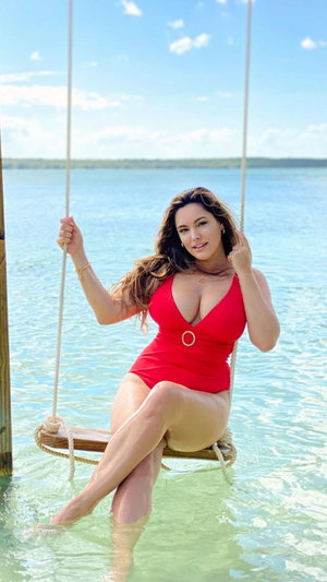 Woman in red one-piece swimsuit sitting on swing over water, legs in clear water.