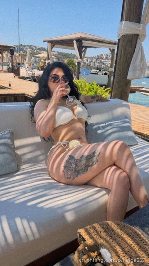Woman in white bikini lounging on outdoor sofa by water, drinking wine, with large thigh tattoo.