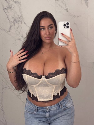 Woman with long dark hair in lace corset and jeans, posing in front of marble wall.