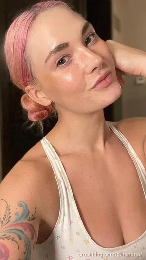 Woman with pink hair and tattoo, wearing a white tank top, posing indoors.