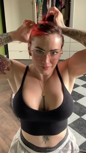 Woman with tattoos and red hair in black crop top, adjusting hair in room with checkered floor.