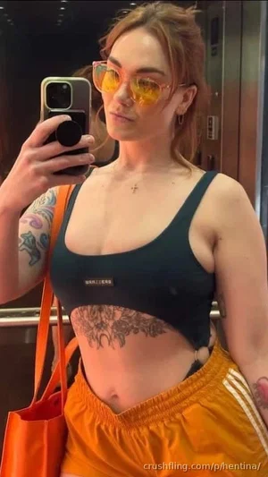 Woman with tattoos in black sports bra and orange shorts, taking mirror selfie.