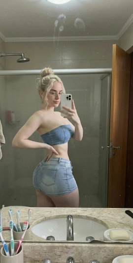 Blonde woman in denim top and shorts taking mirror selfie in bathroom.