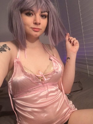 Woman with purple wig in pink satin lingerie, sitting indoors with tattoos visible.