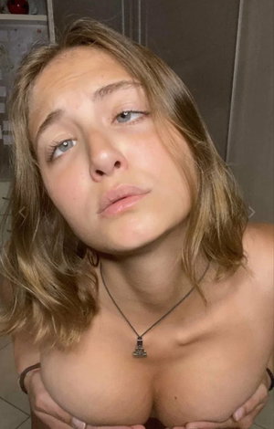 Blonde woman with necklace posing topless indoors.