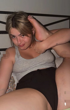Blonde woman in grey tank top and black shorts on bed.