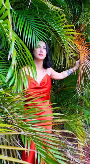 Woman in red dress among palm leaves outdoors.