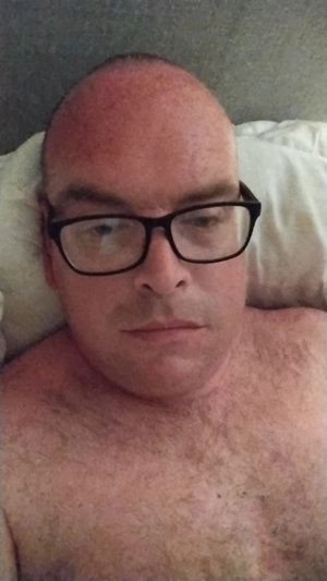 Bald man with glasses lying shirtless on pillow.