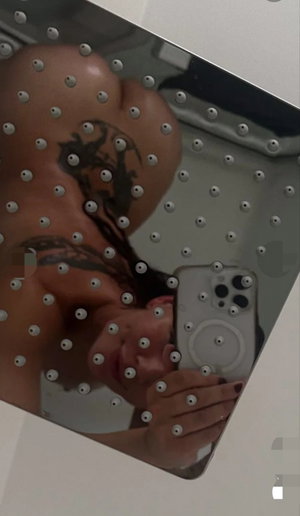 Woman with tattoos taking mirror selfie, bare back and shoulders visible, holding phone.
