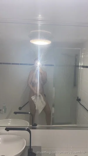 Woman standing in bathroom holding towel, partially covering body, mirror selfie.