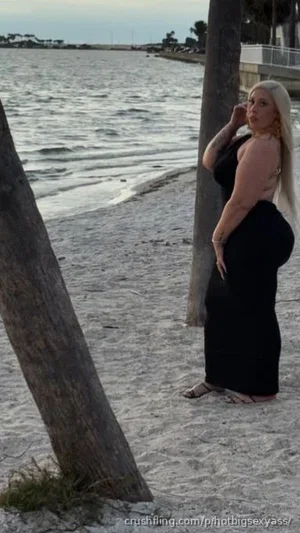 Woman with long blonde hair in black dress standing on beach near palm trees.