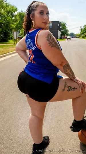 Woman in blue basketball jersey and black shorts posing outdoors on a street with tattoos visible.