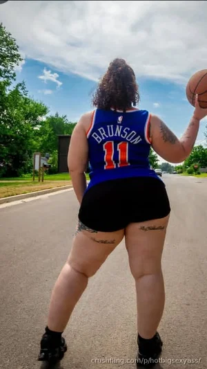 Woman in basketball jersey and black shorts holding a basketball outdoors on a road.