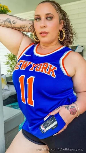 Woman in blue New York basketball jersey and black shorts, posing outdoors with tattoos visible.