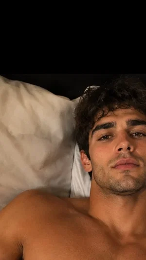 Shirtless man with curly hair lying on a white pillow in a dimly lit room.