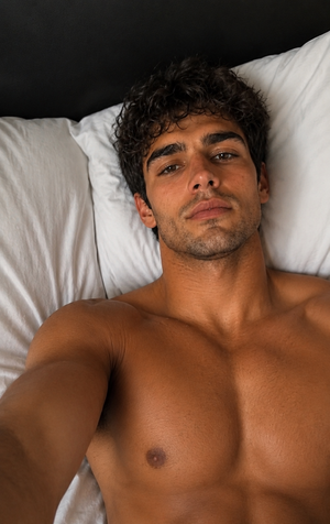 Shirtless man with curly hair lying on white pillows in a bedroom.