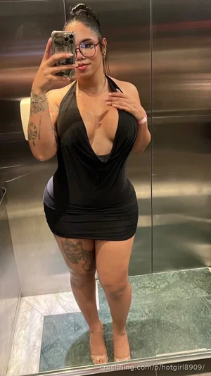 Woman in black halter dress with tattoos, taking mirror selfie in elevator.