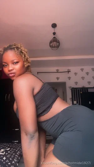 Woman with curly hair in dark top and shorts posing indoors.
