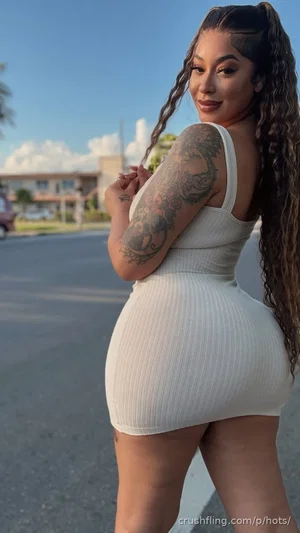 Woman with long curly hair and arm tattoo in white ribbed dress, standing outdoors on street.