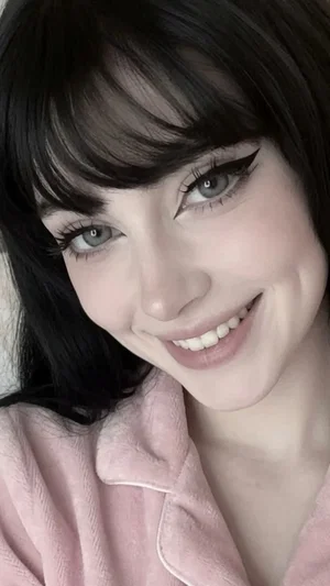 Woman with dark hair and blue eyes wearing pink pajamas, smiling in a close-up selfie.