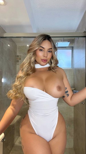 Woman with long wavy hair in white one-piece swimsuit posing in tiled bathroom.