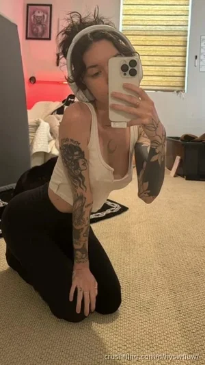 Woman with tattoos in white tank top and black pants, kneeling on carpet in bedroom.