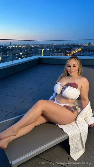 Woman in white lingerie sits on rooftop ledge with city skyline at dusk.