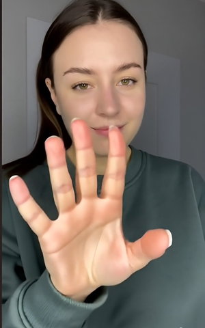 Brunette woman in green sweatshirt waving hand indoors.