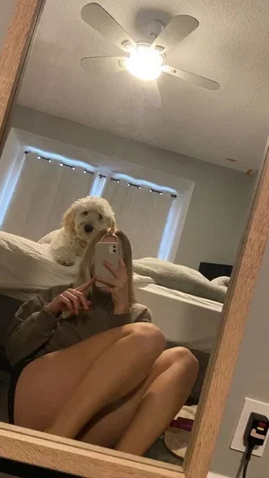 Woman in olive top and black underwear takes mirror selfie in bedroom with dog on bed.