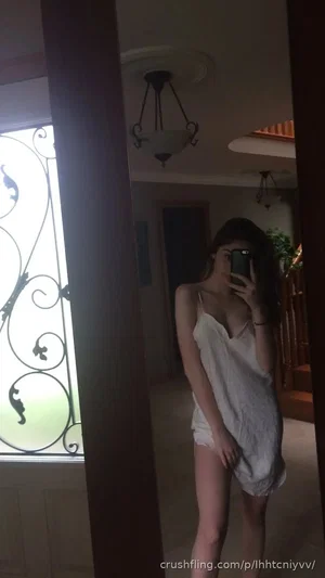 Woman in white slip dress taking mirror selfie in dimly lit indoor space.