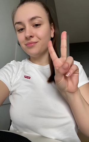 Slim woman in white t-shirt, ponytail, peace sign indoors.