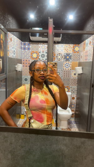 Woman with braids and glasses in tie-dye top taking bathroom selfie.