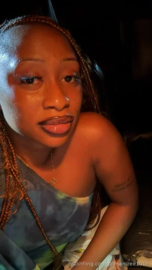 Woman with braided hair in a tie-dye top, posing indoors in dim lighting.