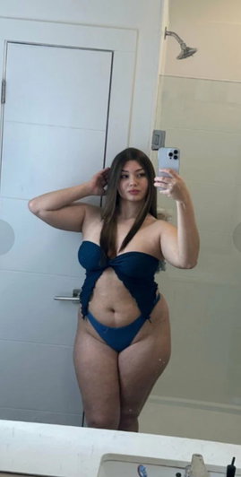 Curvy woman with long brown hair in blue outfit taking mirror selfie.