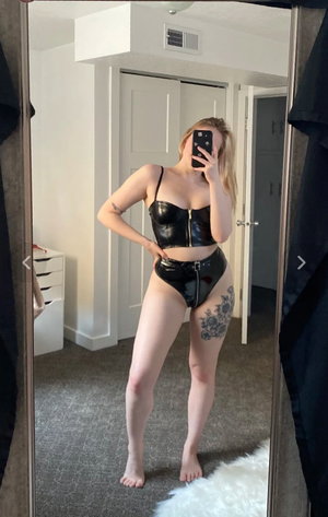 Blonde woman in black shiny outfit taking mirror selfie indoors.