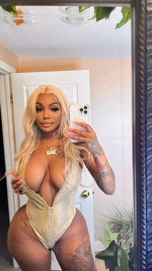 Woman with long blonde hair and tattoos wears sheer lace bodysuit, taking mirror selfie in bedroom.