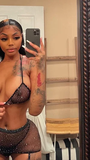 Woman in black mesh lingerie with tattoos takes mirror selfie indoors.