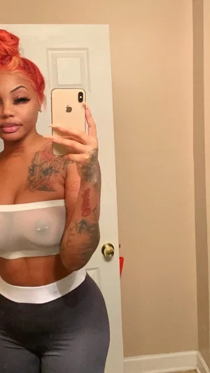 Woman with red hair and tattoos wears sheer top and dark leggings, taking mirror selfie indoors.