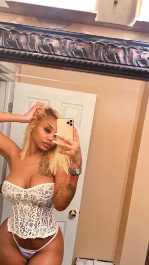 Woman with blonde hair and tattoos wears white lace corset and underwear, taking mirror selfie in