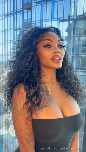 Woman with curly black hair and tattoos wearing a black off-shoulder top, standing by a window with
