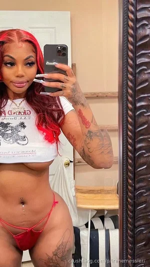 Woman with red hair and tattoos wears white crop top and red thong, taking mirror selfie in bedroom.