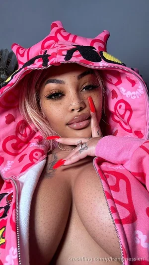 Woman with pink hair and freckles in a pink patterned hoodie, posing indoors with hand near face.