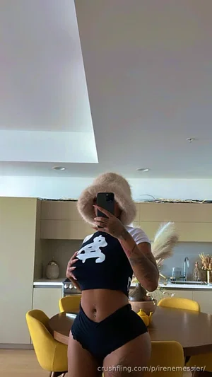 Woman in black crop top and underwear, holding phone, in modern kitchen with yellow chairs.