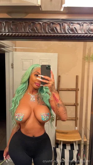 Woman with green hair and tattoos, wearing black leggings and jeweled pasties, taking mirror selfie